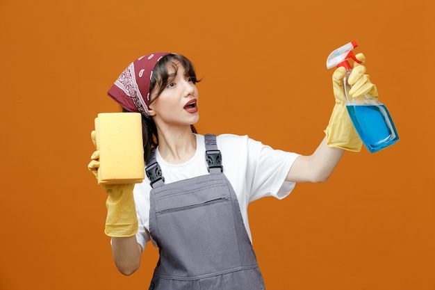 Foto colpito la giovane donna che indossa guanti di gomma uniformi e bandana che tiene la spugna in aria che si allunga il detergente guardando il lato finge di pulire qualcosa di isolato su sfondo arancione