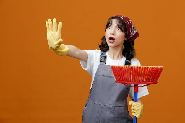 Impressed young female cleaner wearing uniform rubber gloves and bandana holding squeegee mop stretching hand out looking at side showing stop gesture isolated on orange background