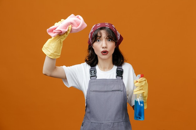 Ha impressionato la giovane donna che indossa guanti di gomma uniformi e bandana che solleva un panno per spolverare tenendo il detergente guardando la fotocamera isolata su sfondo arancione