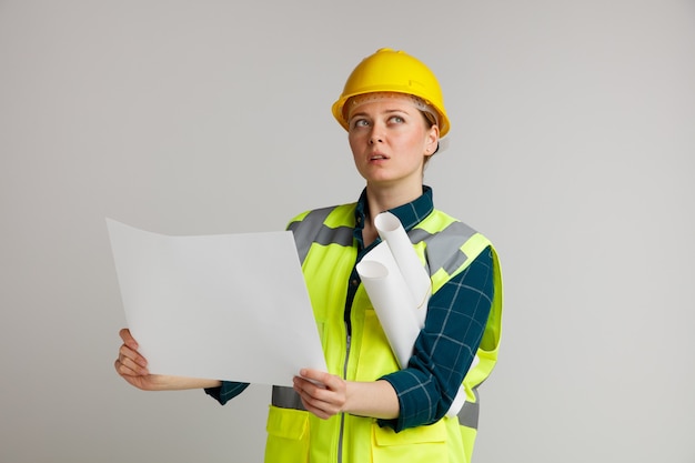 Impressionato giovane operaio edile femminile che indossa il casco di sicurezza e giubbotto di sicurezza che tiene carta e altri sotto il braccio alzando lo sguardo