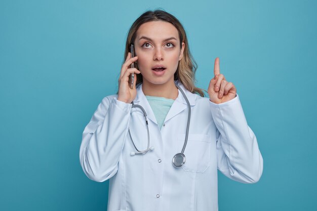 Impressed young female doctor wearing medical robe and stethoscope around neck looking and pointing up 