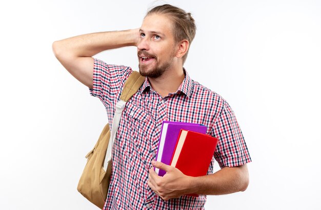 Impressed young guy student wearing backpack holding books putting hand on head isolated on white wall