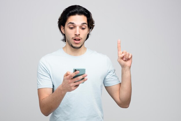 impressed young handsome man holding and looking at mobile phone pointing up isolated on white background