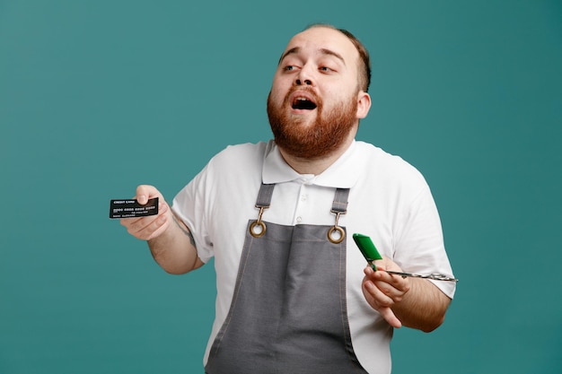 Impressed young male barber wearing white shirt and barber apron looking at side holding credit card scissors and comb isolated on blue background