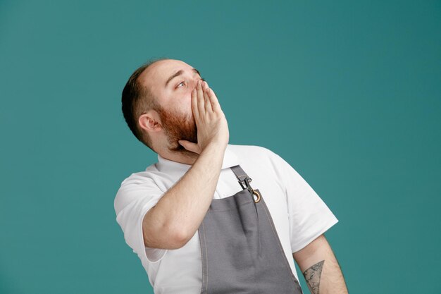 Impressed young male barber wearing white shirt and barber apron looking up showing whisper gesture isolated on blue background