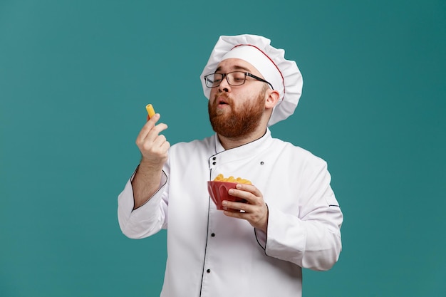 Impressionato giovane chef maschio che indossa occhiali uniformi e cappuccio che tiene una ciotola di maccheroni e un maccheroni in un'altra mano guardando i maccheroni isolati su sfondo blu