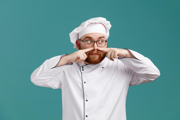 Impressed young male chef wearing glasses uniform and cap looking at side pointing fingers at nose isolated on blue background