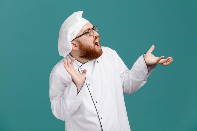 Impressed young male chef wearing glasses uniform and cap looking at side showing empty hands isolated on blue background