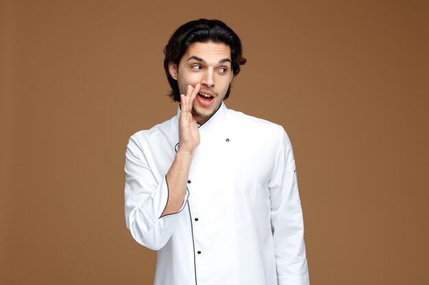 impressed young male chef wearing uniform keeping hand near mouth looking at side whispering isolated on brown background