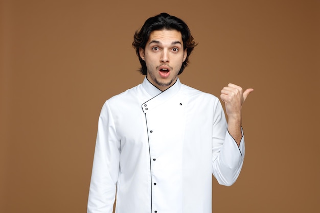 impressed young male chef wearing uniform looking at camera pointing to side isolated on brown background