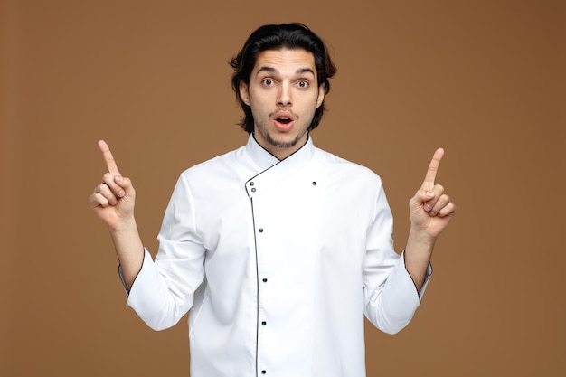 impressed young male chef wearing uniform looking at camera pointing up isolated on brown background