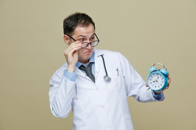 Impressed young male doctor wearing glasses medical robe and stethoscope around neck holding alarm clock grabbing glasses looking at alarm clock from behind glasses isolated on purple background