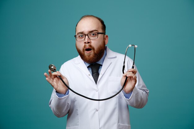 Impressed young male doctor wearing medical coat and glasses holding stethoscope looking at camera isolated on blue background