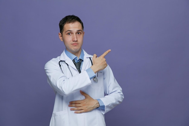 Impressionato giovane medico maschio che indossa un abito medico e uno stetoscopio intorno al collo tenendo la mano sulla pancia guardando la fotocamera che punta al lato isolato su sfondo viola con spazio per la copia