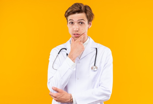 Impressed young male doctor wearing medical robe with stethoscope grabbed chin isolated on orange wall