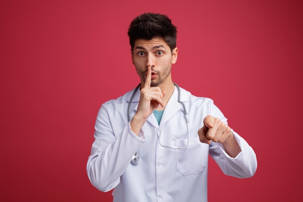 Impressed young male doctor wearing medical uniform and stethoscope around his neck looking and pointing at camera showing silence gesture isolated on red background