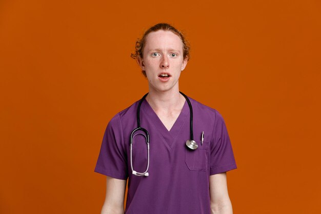 impressed young male doctor wearing uniform with stethoscope isolated on orange background