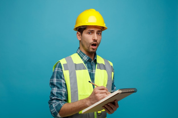 Impressed young male engineer wearing safety helmet and uniform holding pencil and note pad looking at camera isolated on blue background