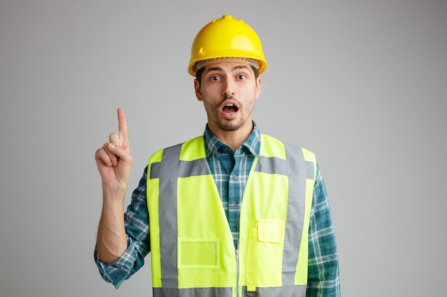 Impressed young male engineer wearing safety helmet and uniform looking at camera pointing finger up isolated on white background