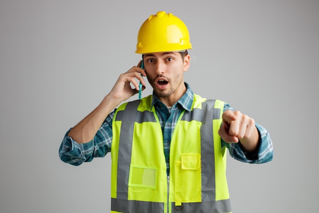Впечатленный молодой инженер-мужчина в защитном шлеме и униформе смотрит и указывает на камеру во время разговора по телефону на белом фоне