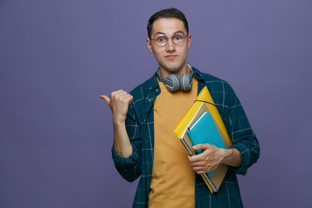 Impressed young male student wearing glasses headphones around neck holding note pad folder note book looking at camera pointing to side isolated on purple background with copy space