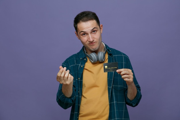 Impressed young male student wearing headphones around neck looking at camera showing credit card and money gesture isolated on purple background