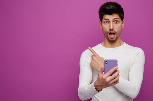 Impressed young man holding mobile phone looking at camera pointing to side isolated on purple background with copy space