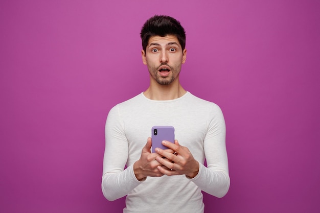 Impressed young man holding mobile phone with both hands looking at camera isolated on purple background