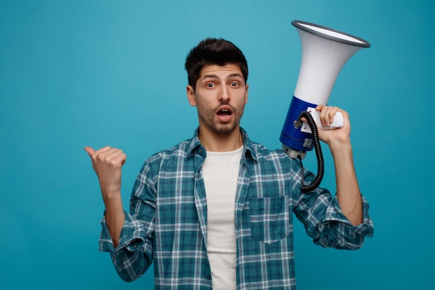 Impressed young man holding speaker looking at camera pointing to side isolated on blue background