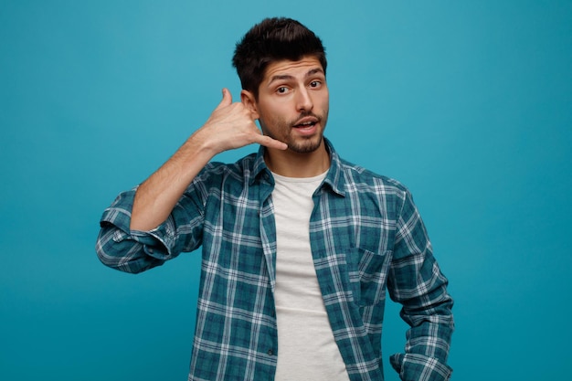 Impressed young man looking at camera showing call me gesture isolated on blue background