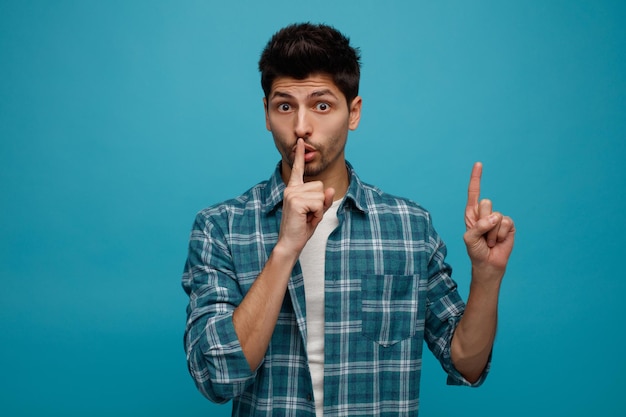 Impressed young man looking at camera showing silence gesture pointing up isolated on blue background