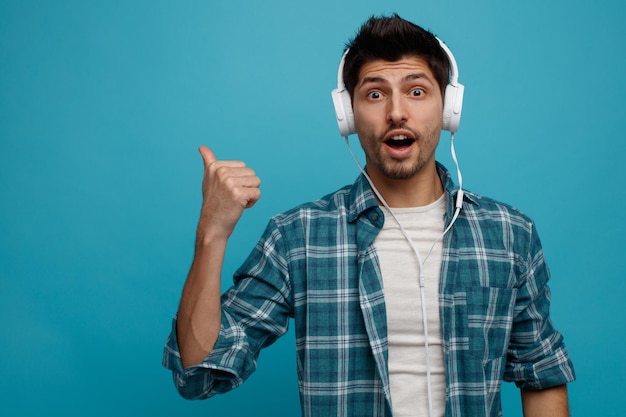 Impressed young man wearing headphones looking at camera pointing to side isolated on blue background