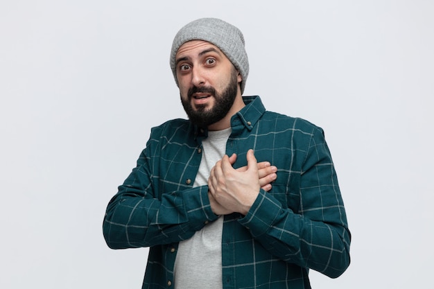 Impressed young man wearing winter hat keeping hands on chest looking at camera isolated on white background