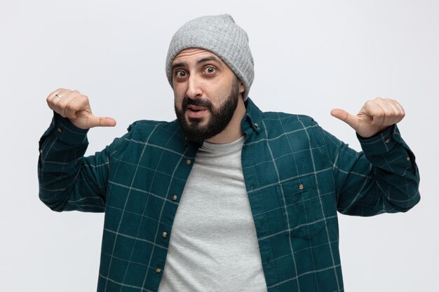 Impressed young man wearing winter hat looking at camera pointing at his front isolated on white background