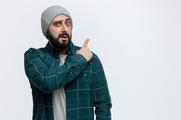 Impressed young man wearing winter hat looking at camera pointing up isolated on white background with copy space