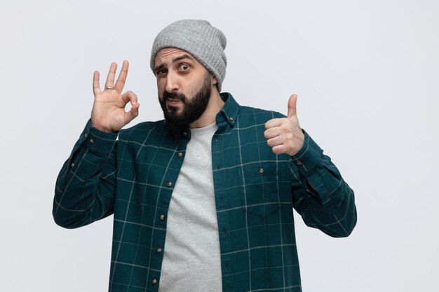 Impressed young man wearing winter hat looking at camera showing ok sign and thumb up isolated on white background