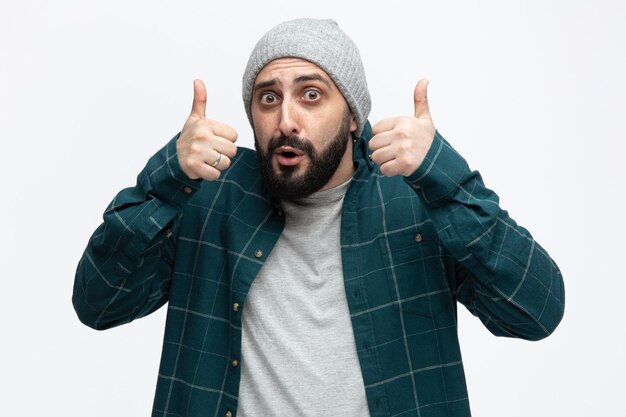 Impressed young man wearing winter hat looking at camera showing thumbs up isolated on white background