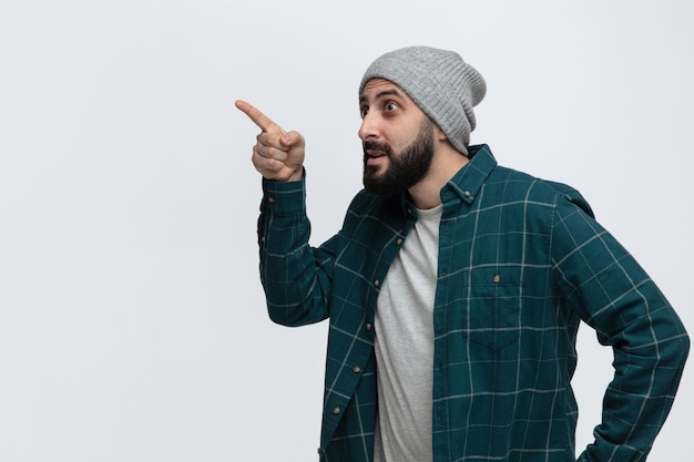 Impressed young man wearing winter hat looking and pointing at side isolated on white background with copy space