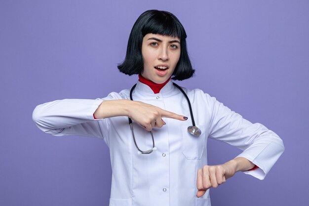 Impressionata giovane bella ragazza caucasica in uniforme da medico con uno stetoscopio che punta alla sua mano