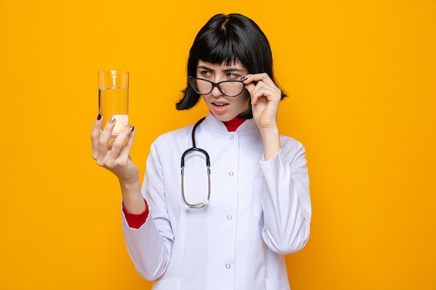 Impressionata giovane bella ragazza caucasica con occhiali ottici che indossa un'uniforme da medico con uno stetoscopio che tiene in mano un bicchiere d'acqua