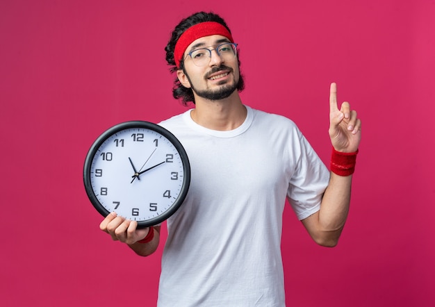 Impressed young sporty man wearing headband with wristband holding wall clock points at up 