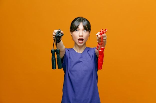 impressed young sporty woman wearing tshirt looking at camera stretching jumping ropes out towards camera isolated on orange background