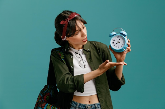 Impressed young student girl wearing bandana and backpack showing alarm clock looking and pointing with hand at it with glasses on her blouse isolated on blue background