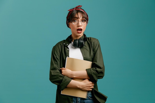 Impressed young student girl wearing bandana glasses and headphones around neck holding and hugging large note pad looking at camera isolated on blue background