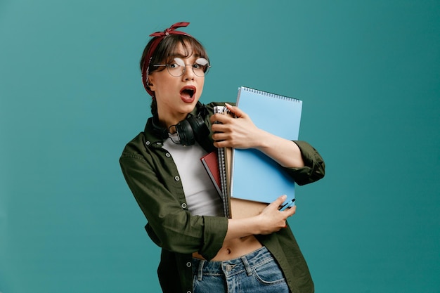 Impressed young student girl wearing bandana glasses and headphones around neck holding pen and large note pads looking at camera isolated on blue background