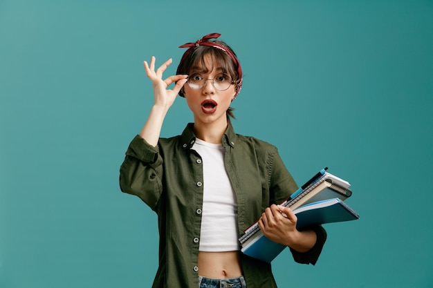 Impressed young student girl wearing bandana glasses holding large note pads with pen grabbing glasses looking at camera isolated on blue background