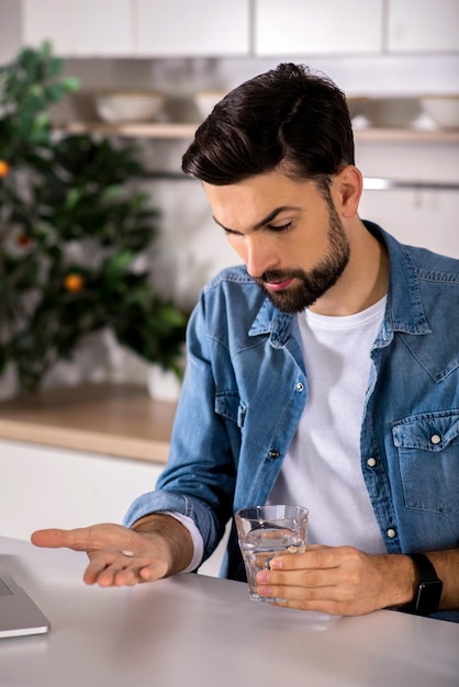 Improve your health Handsome bearded yougn man sitting in the kitchen while taking pills