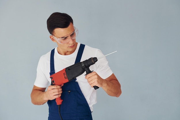 In beschermende brillen en met boor in handen Mannelijke werknemer in blauw uniform staande in studio tegen witte achtergrond