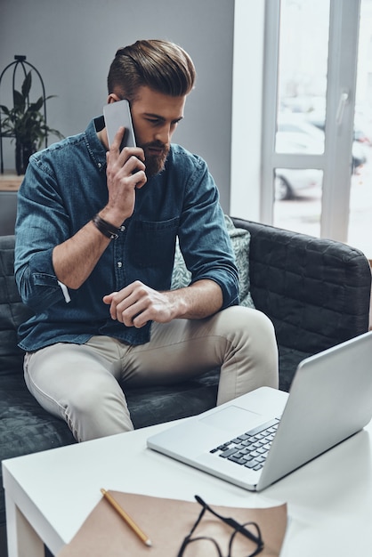 In contact met opdrachtgevers. Doordachte jonge moderne man die op zijn smartphone praat terwijl hij op zijn werkplek zit