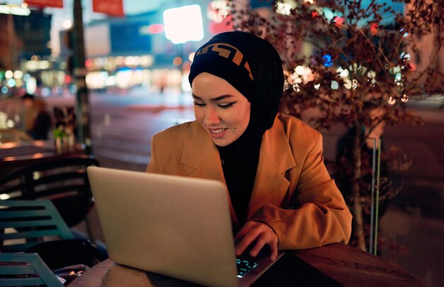 In de betoverende ambiance van een nachtelijk stadsbeeld creëert een in hijab gekleed meisje verdiept in haar laptop